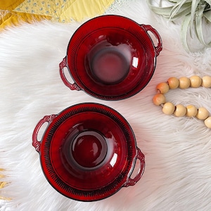 May include: Two red glass bowls with black trim and handles. The bowls are on a white fluffy surface.