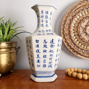 May include: A white ceramic vase with blue Chinese characters and geometric patterns. The vase has a hexagonal shape with a flared top and a blue base. The vase is decorated with Chinese calligraphy.