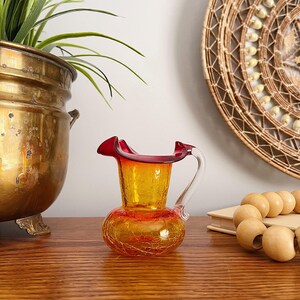 May include: A small, vintage glass pitcher with a crackle finish. The pitcher features a bulbous base and a flared, ruffled rim in red. The body is yellow and orange, with a clear glass handle. A decorative piece for a home.