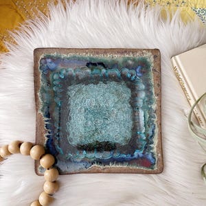 May include: A square ceramic dish featuring a textured, light blue glass center surrounded by a dark blue and brown glazed border. The edges of the dish are slightly raised and uneven, showcasing a reactive glaze finish with deep blue and teal tones.