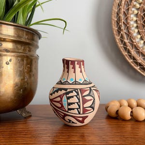 May include: A small, beige ceramic vase with a rounded body and a narrow neck. The vase is adorned with intricate geometric patterns in black, red, and turquoise. The vase is displayed on a wooden surface.