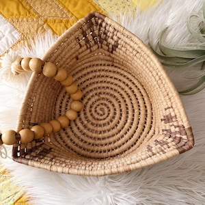 May include: A triangular woven basket featuring a coiled design with light tan and dark brown fibers. The basket includes a decorative handle made of smooth, round, light-colored wooden beads strung on a white cord attached to one corner.