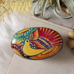 May include: Oval-shaped, hand-painted decorative dish featuring a yellow bird with a blue beak. The bird is surrounded by colorful, stylized feathers in red, teal, and purple. The dish has a blue border and is likely made of ceramic or a similar material.