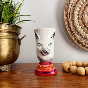 May include: A white ceramic cat-faced vase with blue eyes, black ears, and a red and yellow patterned base. The vase is on a wooden surface, with a brass pot and woven wall decor in the background.