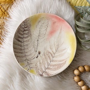 May include: A round, off-white ceramic bowl with a fern leaf design. The bowl features a gradient of yellow and pink hues. The fern design is in shades of brown. The bowl is a decorative piece.