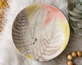 May include: A round, off-white ceramic bowl with a fern leaf design. The bowl features a gradient of yellow and pink hues. The fern design is in shades of brown. The bowl is a decorative piece.