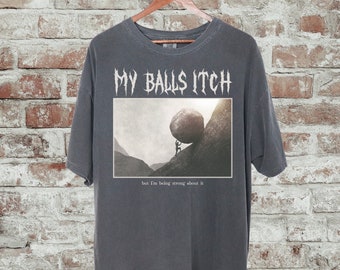 May include: A dark gray t-shirt with a graphic print of a person pushing a large rock uphill. The text "MY BALLS ITCH" is printed in white above the graphic. The text "but I'm being strong about it" is printed in white below the graphic.