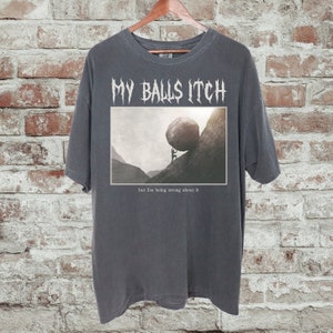 May include: A dark gray t-shirt with a graphic print of a person pushing a large rock uphill. The text "MY BALLS ITCH" is printed in white above the graphic. The text "but I'm being strong about it" is printed in white below the graphic.
