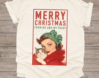May include: Cream-coloured t-shirt with a festive graphic. The design shows a woman in a green hat and red coat cuddling a cat, with the text "MERRY CHRISTMAS FROM ME AND MY PUSSY" in red and green.