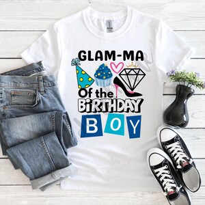Pode incluir: Camiseta branca com o texto "GLAM-MA Of the BIRTHDAY BOY" em preto e azul. O design inclui um chapéu de festa, um cupcake, um coração, um diamante e um salto alto. Uma calça jeans azul e tênis pretos também estão na imagem.