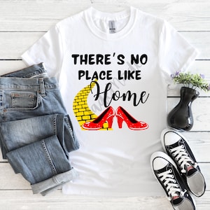 Puede incluir: Camiseta blanca con la frase "THERE'S NO PLACE LIKE HOME" y una ilustración del camino de baldosas amarillas y zapatos rojos. La imagen también incluye unos vaqueros azules y zapatillas deportivas blancas y negras.