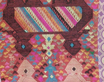 May include: A red fabric with intricate woven patterns in various colours, including black, white, yellow, blue, and pink. The fabric features geometric designs and stylised animal figures.