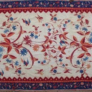 May include: A white fabric with a red, blue, and orange floral pattern. The fabric has a border with a repeating pattern of red, blue, and white.