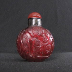 Könnte beinhalten: Eine rote, runde Schnupftabakflasche aus Glas mit einem silberfarbenen Metallhals und einem roten Verschluss. Die Flasche ist mit detaillierten Figuren und floralen Designs geschnitzt. Ein dekorativer Artikel.