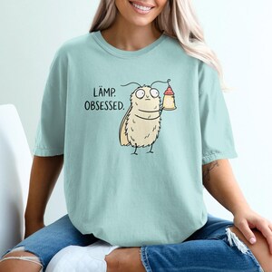 May include: A light blue t-shirt featuring a cartoon illustration of a fuzzy moth holding a small lamp. The text "LÄMP. OBSESSED." is printed in black letters above the moth. The shirt has a crew neckline and short sleeves with a relaxed fit.