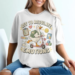 Peut inclure: Un t-shirt blanc avec un motif représentant un canard blanc au bec et aux pattes orange sur une trottinette rose. Le canard porte un sac à dos vert. Le texte indique OFF TO REGULATE EMOTIONS. On y voit aussi un presse-papier, un smiley, des étoiles, des empreintes de mains et des blocs colorés.