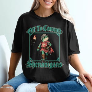 Può includere: Una t-shirt nera con la grafica di una rana verde vestita da cavaliere medievale con elmo, tunica rossa e spada. Il testo "Off To Commit" appare sopra la rana e "SHENANIGANS" sotto, il tutto incorniciato da un bordo color verde acqua.