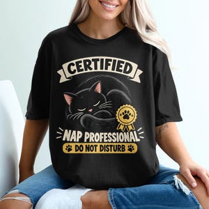 May include: A black short-sleeve t-shirt featuring a graphic of a sleeping black cat. The text reads CERTIFIED NAP PROFESSIONAL DO NOT DISTURB. A yellow award ribbon icon with a paw print is positioned next to the cat. The design uses cream and yellow.