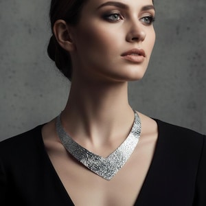 May include: A silver-colored statement necklace with a textured, organic design. The necklace has a wide, V-shaped pendant and is worn against a black dress. The woman in the image has dark hair and is looking to the side.