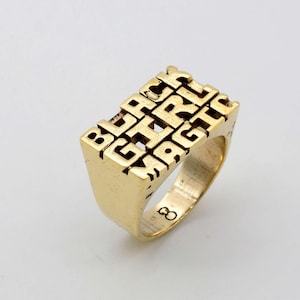 May include: A gold ring with the words "BLACK GIRL MAGIC" spelled out in raised letters.