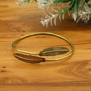 May include: A gold-colored arm cuff bracelet with two decorative feather accents. The bracelet is open and adjustable, with the feather details positioned on the ends. The feathers have a detailed, textured design.