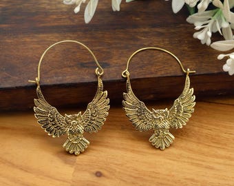 May include: A pair of gold-colored hoop earrings. Each earring features a detailed owl design with outstretched wings. The earrings are made of a shiny, metallic material and are displayed on a wooden surface.