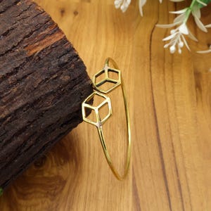 May include: A gold-colored bangle bracelet with two geometric cube-shaped accents. The bracelet is thin and circular, with the cube shapes positioned near the top. The bracelet is displayed on a wooden surface.