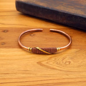 May include: Copper cuff bracelet with a woven center section and gold-colored accents. The open-ended bracelet sits on a wooden surface. The bracelet's design features a combination of copper and gold tones.
