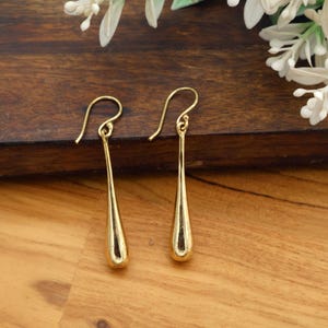 May include: A pair of gold-colored teardrop earrings. The earrings have a simple, elegant design with a curved hook at the top and a smooth, elongated teardrop shape. The earrings are displayed on a wooden surface.