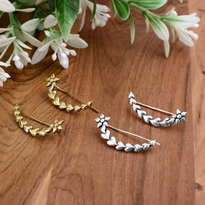 May include: Four decorative hair pins. Two are gold-toned, and two are silver-toned. Each pin features a floral design with a curved bar and leaf accents. The pins are arranged on a wooden surface with white flowers in the background.