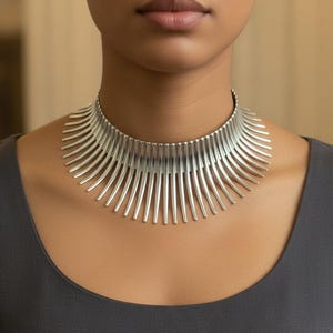May include: A silver-toned collar necklace with a striking, modern design. The necklace features numerous vertical, curved bars that fan out from the neckline. The jewelry is worn against a dark gray top, creating a contrast that highlights the necklace's metallic sheen.