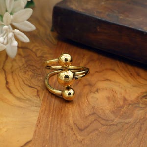 May include: A gold-colored ring with three spherical accents. The ring has a spiral design, with the spheres arranged along the spiral. The ring is displayed on a wooden surface, with a dark wooden box in the background.
