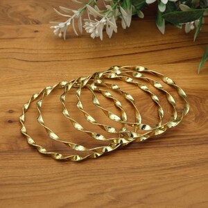 May include: A set of six gold-colored bangles with a twisted rope design. The bangles are arranged on a wooden surface, with white flowers in the background. The bangles have a shiny, metallic finish.