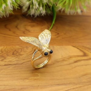 May include: A gold-colored ring in the shape of a fly, with detailed wings and two black gemstone eyes. The ring is set against a wooden surface, showcasing the intricate design and craftsmanship of the jewelry.