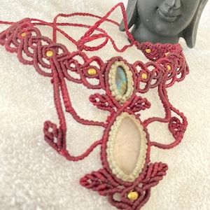 May include: A handmade burgundy macrame necklace with a central oval-shaped rose quartz stone and a smaller oval-shaped labradorite stone. The necklace features gold-colored beads and intricate knotting details, creating a bohemian style.