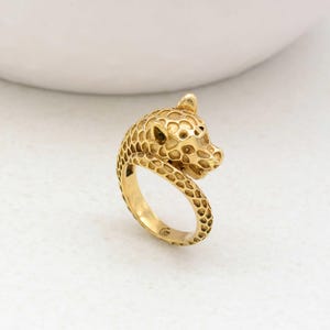 May include: A gold-toned ring featuring a detailed, curled leopard design. The ring is made of metal and has a textured surface.
