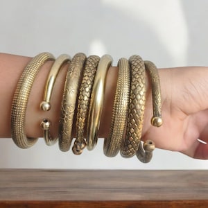 May include: A set of eight gold-toned metal bangles with various textures and patterns. The bangles are stacked on a person's wrist.