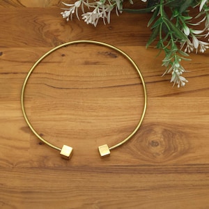 May include: A gold-colored collar necklace with a circular design. The necklace has a simple, elegant style, with a smooth, polished finish. The ends of the necklace feature small, square-shaped accents. The necklace is displayed on a wooden surface.