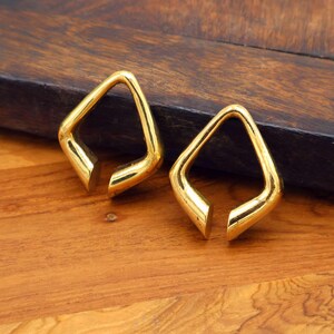 May include: Two gold-colored, open-ended, diamond-shaped earrings. The earrings are displayed on a wooden surface with a dark brown background. The earrings have a polished, reflective surface.
