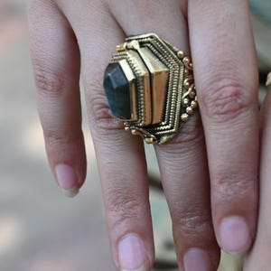 May include: A gold-toned ring with a large, geometric design. The ring features a central, dark-colored stone set within a detailed, ornate frame. The ring's design includes hexagonal and rectangular shapes, with intricate detailing.