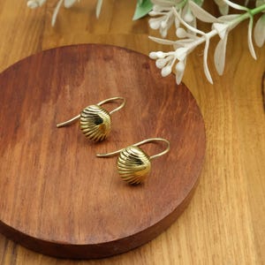 May include: A pair of gold-colored earrings with a spherical, ridged design. The earrings are displayed on a round, wooden surface, with white flowers in the background. The earrings have a hook closure.