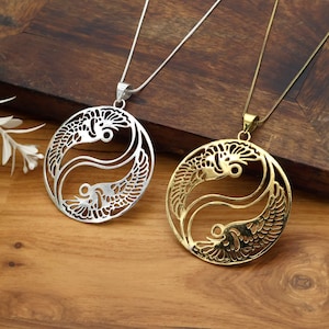 May include: Two pendant necklaces with a yin-yang design featuring stylized fish. One is silver-colored, the other gold-colored. Both pendants hang from delicate chains. The necklaces rest on a wooden surface.