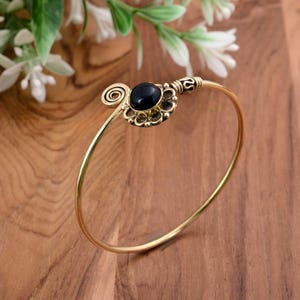 May include: A gold-colored bangle bracelet with a black gemstone center. The bracelet features an ornate design around the gemstone and a spiral detail on one end. The background includes white flowers and a wooden surface.