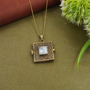 May include: A square, antique gold-colored pendant necklace with a square, light-colored stone in the center. The pendant has an ornate, filigree design and hangs from a delicate gold chain. The necklace is displayed on a green surface.