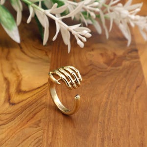 May include: A gold-colored ring featuring a skeletal hand design. The ring's hand is open, with the fingers slightly curved. The ring is set against a wooden surface, with white floral accents in the background.