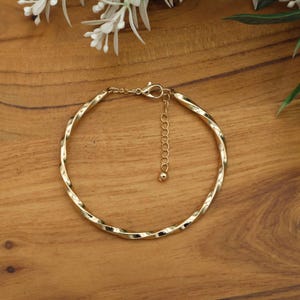 May include: A gold-colored bracelet with a twisted design. The bracelet has a clasp closure and an adjustable chain. The jewelry is displayed on a wooden surface with white flowers and green leaves in the background.