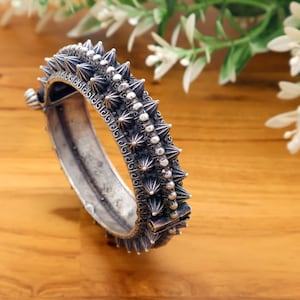 May include: A silver-toned bangle bracelet with a spiked design. The bracelet features a series of conical spikes and small round beads along its surface. The metal has an aged appearance, and the bracelet is set against a wooden background.