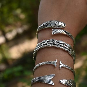 May include: Silver-toned cuff bracelets with fish designs. The bracelets feature detailed scales and open-ended designs, with some depicting fish heads and tails. The bracelets are worn on an arm, showcasing the intricate metalwork.