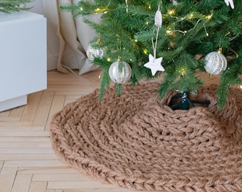 May include: A round, chunky knit Christmas tree skirt in a solid tan color. The skirt features a thick, braided yarn texture and is positioned at the base of a green Christmas tree decorated with white ornaments and warm white string lights.