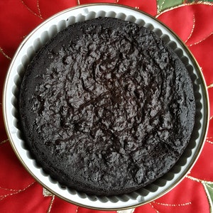 May include: A dark chocolate cake in a white baking pan with a fluted edge. The cake is round and has a slightly cracked surface.